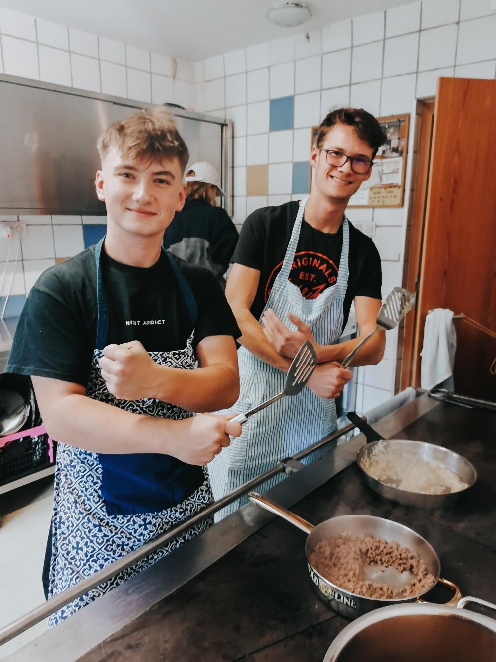 Zwei junge Männer mit Schürze, welche in einer Küche stehen und kochen.